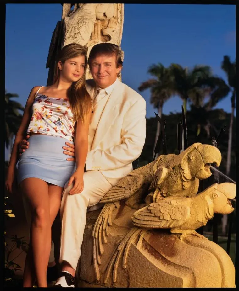Ivanka and Trump are pictured outdoors in this portrait. Ivanka is seated on a stone sculpture featuring intricately carved parrots. She is wearing a sleeveless floral top and a light blue skirt, her long hair cascading over her shoulders. Trump is beside her, dressed in a crisp white suit, exuding a formal yet relaxed demeanor. The background features lush palm trees and clear skies, indicating a sunny day, enhancing the warm and vibrant ambiance of the scene. The lighting is soft, casting a pleasant glow on their faces. The sculpture adds an element of artistic detail, hinting at an opulent setting. Both appear poised and content, with a serene atmosphere enveloping the scene.