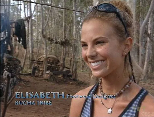 Elisabeth Hasselbeck is shown in an outdoor setting, likely part of a reality TV show. She is smiling, indicating a positive mood, and is wearing a sleeveless top and a beaded necklace. The background consists of a forested area, suggesting a survival or wilderness theme. The text on the screen identifies her as "ELISABETH," a footwear designer from the "KUCHA TRIBE," linking her to the show "Survivor" on CBS. The image captures a candid moment typical of interview segments in reality TV shows, providing insight into the contestant's role and personality on the show. The lighting is natural, emphasizing the outdoor environment and the raw, authentic atmosphere typical of "Survivor."