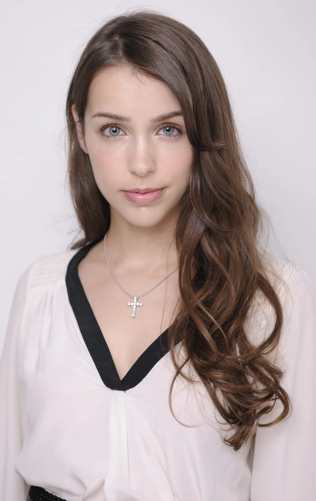 Stefanie Joosten is depicted in this portrait image. The setting is a simple, well-lit indoor studio with a plain background, emphasizing the subject. Stefanie Joosten has long, wavy brown hair cascading over her shoulders, and she is wearing a light-colored blouse with a distinct black trim at the neckline. A notable accessory is a cross-shaped necklace adorned with small stones, giving a touch of elegance. Her expression is calm and composed, with a gentle smile that conveys a serene and approachable mood. The lighting is soft, highlighting her features and creating a natural look. The image is a close-up shot, focusing on her face and upper torso, ensuring that her style and accessories stand out clearly. There is no visible text or branding in the image, maintaining focus solely on Stefanie Joosten.