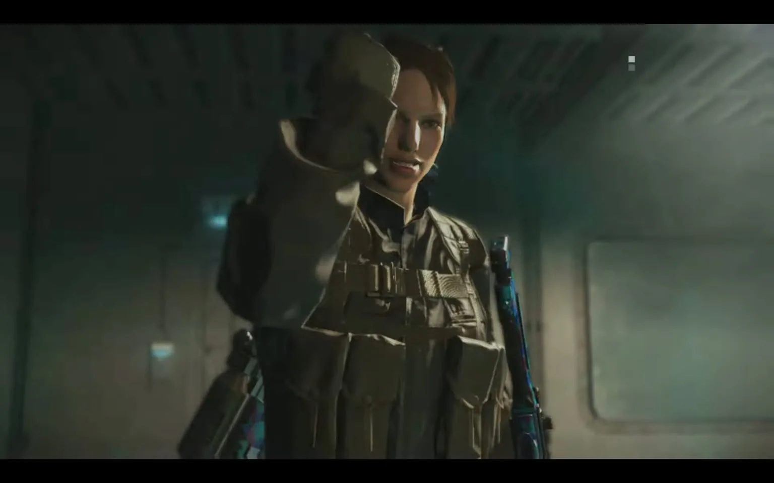 The image shows a character from "Modern Warfare III" inside a dimly lit, industrial interior, likely a vehicle or a ship. The atmosphere is intense, with muted colors and a focus on the character's determined expression. She is dressed in tactical military gear, including a vest with utility pouches and a long-sleeved uniform. The character is holding up her arm in a commanding gesture, suggesting urgency or resolve. Her weapon, which appears to be a rifle, is slung across her back, indicating readiness for action. The lighting is subdued, casting shadows that enhance the serious tone of the scene. The setting appears to be a confined space, adding to the tension and focus on the character's actions.