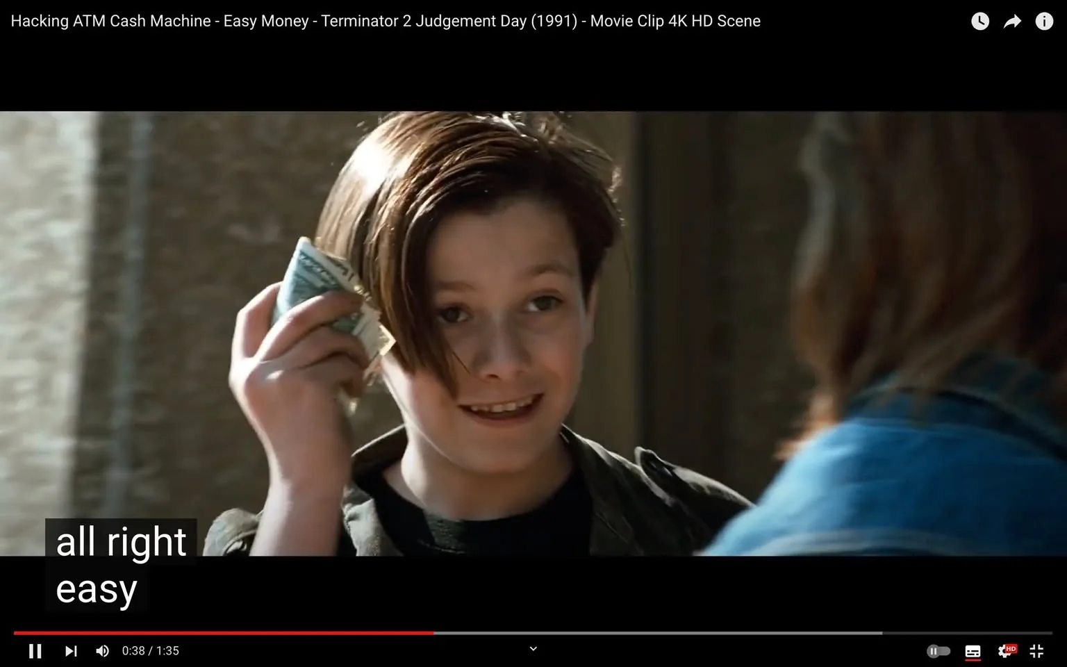 The video frame shows a scene from "Terminator 2 Judgement Day," capturing an outdoor setting during daytime. In the foreground, a young character is seen holding a stack of cash beside their face, showcasing a confident and pleased expression. The character's hair is styled in a classic 90s fashion, with a side part and medium length, giving a youthful appearance. They are wearing a dark jacket over a black shirt, indicative of a casual look. The lighting suggests clear daylight, casting natural shadows and highlighting the character’s features. In the background, the setting appears to be an urban area, though details are blurred, keeping the focus on the character.
The video is titled "Hacking ATM Cash Machine - Easy Money - Terminator 2 Judgement Day (1991) - Movie Clip 4K HD Scene," suggesting a scene involving technology or hacking. The subtitles at the bottom of the frame read "all right easy," possibly indicating dialogue or commentary related to the action in the scene. The setting and attire suggest a typical environment for a young character in a 1990s action film.