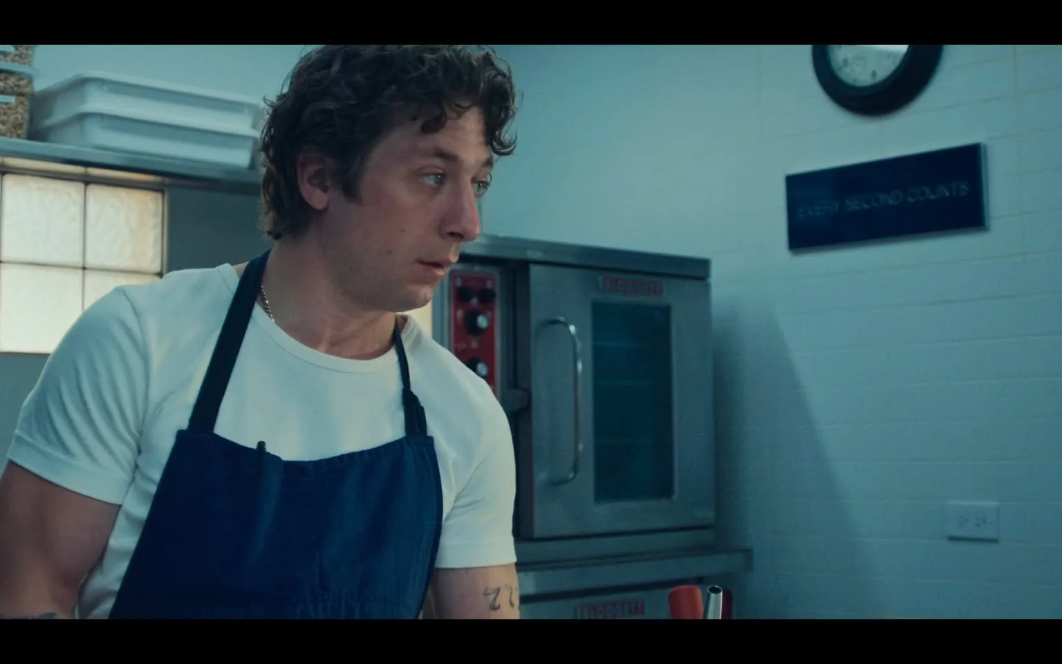 In this scene from "The Bear," Jeremy Allen White is seen in what appears to be a professional kitchen. He is wearing a white t-shirt with a navy blue apron, suggesting his role as a chef or someone involved in culinary work. The setting is well-lit, with a background that includes a commercial oven and a wall clock, adding to the realistic kitchen environment. A sign on the wall reads "EVERY SECOND COUNTS," indicating the high-pressure nature of the setting. Jeremy's expression is focused and intense, reflecting the demanding atmosphere of a busy kitchen. The color palette is cool with shades of blue and white dominating the scene, creating a clean and efficient ambiance. His posture is attentive, possibly indicating he is listening or preparing to act swiftly, embodying the urgency suggested by the environment.