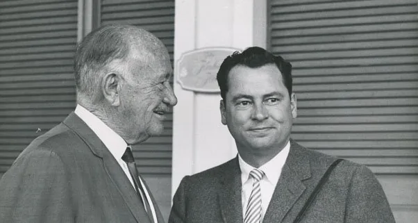 This black and white photograph shows two men standing outdoors, engaged in conversation. The man on the left, who the hint suggests could be Arthur Sulzberger, is an older gentleman with a receding hairline and a distinguished, weathered face. He is wearing a suit and tie, exuding a formal and professional demeanor. The man on the right, possibly Bernie Sanders as per the hint, appears younger, with neatly combed dark hair and a confident expression. He is also dressed formally, wearing a suit with a tie, and has a more relaxed posture as he gazes slightly to the side.
The background consists of a structure with closed shutters, suggesting an outdoor setting, perhaps at a formal event or gathering. The lighting is natural, highlighting the details of their facial expressions and attire. The atmosphere seems congenial, with the two men appearing to be in a moment of casual interaction, possibly discussing business or a related topic. The image captures a moment of historical significance, possibly connected to their roles or contributions in their respective fields.