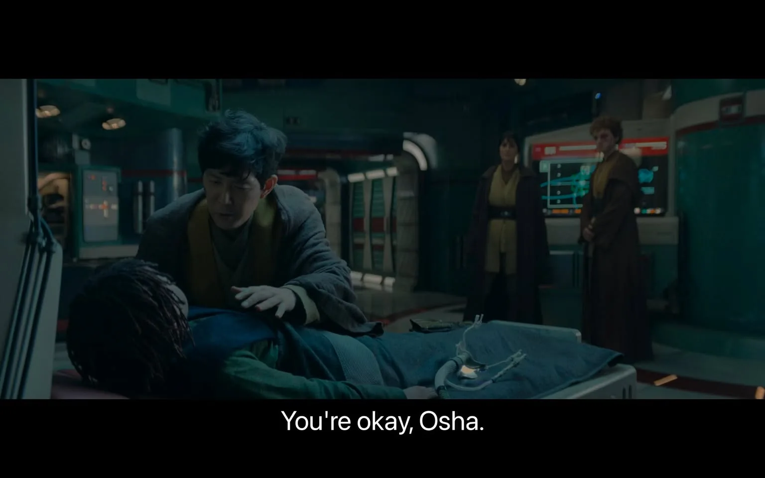The image depicts a scene set in a futuristic medical bay. In the foreground, a person with short dark hair, dressed in a robe-like outfit, is attending to someone lying on a medical table, referred to as "Osha" by the subtitle. Osha appears to be resting with medical equipment connected to them. The setting is illuminated with soft, bluish lighting, adding to the sci-fi ambiance. In the background, two individuals stand near a display screen, which shows complex graphics, possibly related to medical monitoring. They are also wearing similar robe-like attire. The room has a sleek, metallic design with high-tech elements, reinforcing the sci-fi theme. The overall atmosphere suggests concern and care within a technologically advanced environment.
