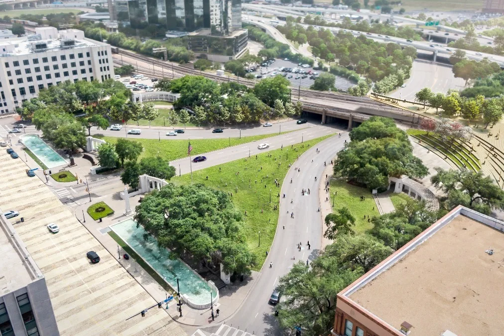 The image depicts an aerial view of an urban park area intersected by roads, with a mix of greenery and urban infrastructure. A triangular patch of grass is centrally located, bordered by curved roads on either side. Pedestrians are visible walking along the roads and grassy area, suggesting a leisurely atmosphere. Surrounding the park, a variety of trees provide shade and add to the lush landscape. To the left, there are two long, narrow pools of water, providing a reflective surface that complements the natural setting. Nearby, buildings and office towers can be seen, indicating the park's location within a bustling cityscape. The background features a network of highways and overpasses, showcasing the integration of nature within an urban environment. The overall lighting is bright and clear, suggesting a sunny day.