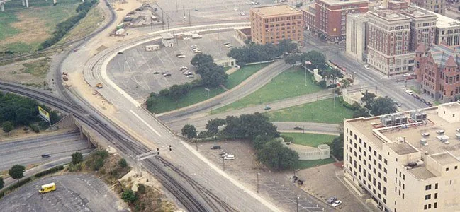 This aerial photograph captures Dealey Plaza, a historically significant area in Dallas, Texas. The image showcases a distinctive layout with curved roads and lush green spaces bordered by urban infrastructure. To the right, notable buildings with varied architectural styles are visible, including a red-bricked structure with a pointed roof and other multi-story edifices. The roadways, primarily concrete, create a network of routes converging at the plaza, with some areas visibly marked for parking. The railway tracks run parallel to the roads, adding to the complexity of the city's layout. The color palette is natural, with greens from the grass and trees contrasting against the muted grays and browns of the urban structures. Lighting appears natural, likely captured during daylight hours, giving the image a clear and detailed view of the area. In the background, further cityscape details fade into the distance, providing a sense of a bustling urban environment.