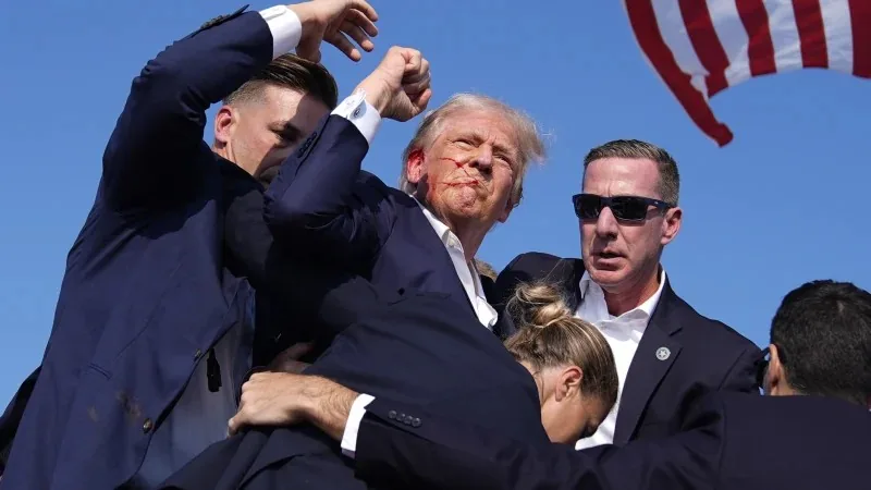 Donald Trump is at the center of the image, surrounded by a group of men who appear to be security personnel. Among them is Sean Curran. Trump is standing with a raised fist, his face appearing slightly strained, suggesting a moment of tension or disruption. He is wearing a dark suit with a white shirt, and his hair is characteristically styled. The security personnel surrounding him are also dressed in suits, and one of them is wearing sunglasses. The background features a clear blue sky with part of an American flag visible, indicating an outdoor location, possibly at a public or political event. The mood is intense, with the security team appearing focused and vigilant, suggesting they are actively responding to a situation. The composition captures a dynamic moment filled with movement and urgency, with the flag adding a patriotic element to the scene.