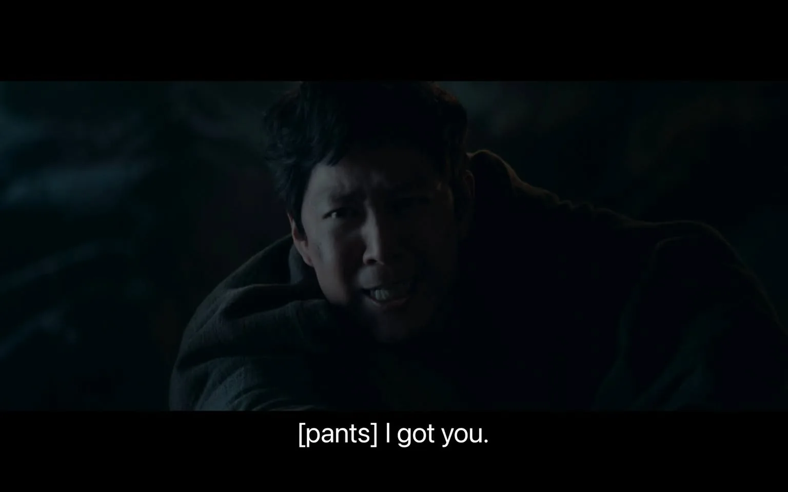 The image shows a dramatic and tense scene with low, moody lighting, suggesting it takes place at night or in a dimly lit setting. The focal point is a person with an intense expression, indicating urgency or concern. They appear to be mid-action, possibly reaching out or holding onto something or someone. The person's face is highlighted, contrasting with the dark background and their dark clothing, which adds to the scene's dramatic atmosphere. The subtitles at the bottom read "[pants] I got you," implying a moment of tension where the person is likely involved in a physical effort or rescue. The overall composition emphasizes the person's expression and action, creating a sense of immediacy and suspense.