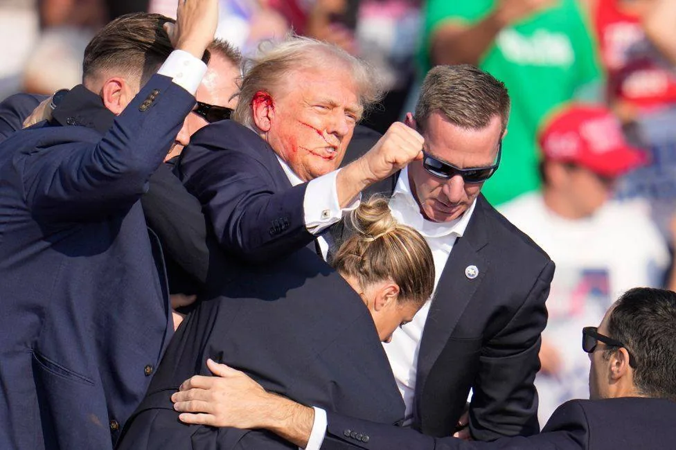 ```
The image captures a moment at a public event where Donald Trump is seen in a group of people, likely security personnel. The focus is on Donald Trump, who appears to be raising his fist while surrounded by several men in suits, suggestive of a security detail. One of the men, identified by the hint as Sean Curran, is wearing sunglasses and appears to be closely guarding Trump. There is an apparent injury on Trump's face, indicated by marks that resemble blood. The background shows a crowd, with some individuals wearing red caps, typically associated with Trump's supporters. The lighting indicates a sunny, outdoor setting, possibly at a rally or public gathering. The atmosphere seems tense, likely due to the visible injury and the presence of security personnel. The image conveys a mix of determination and urgency, highlighted by the expressions and postures of those surrounding Donald Trump.
- Title: "..."