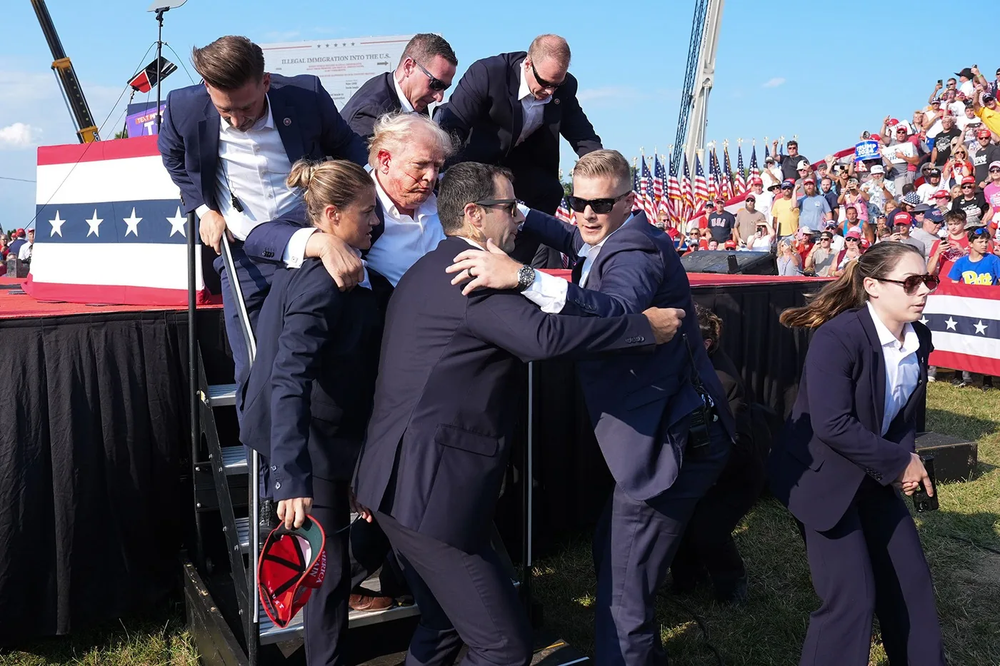 Donald Trump is being assisted by several individuals, likely security personnel, as he descends a set of stairs at an outdoor event. Trump is in the center of the image, looking slightly disheveled with an apparent red mark on the side of his face. He is surrounded by people dressed in suits, indicating a formal and possibly security-related role. The individuals are helping him down the stairs, demonstrating a sense of urgency and care.
In the background, a large crowd is visible, suggesting a public gathering or rally. The crowd is a mix of people, many wearing hats and apparel associated with political events. American flags are prominently displayed, adding a patriotic theme to the scene. The lighting is bright, indicating daytime, with a clear blue sky above. The stairs Trump is descending are part of a stage setup, which is decorated with a banner featuring stars, reinforcing the event's formal and patriotic nature.
