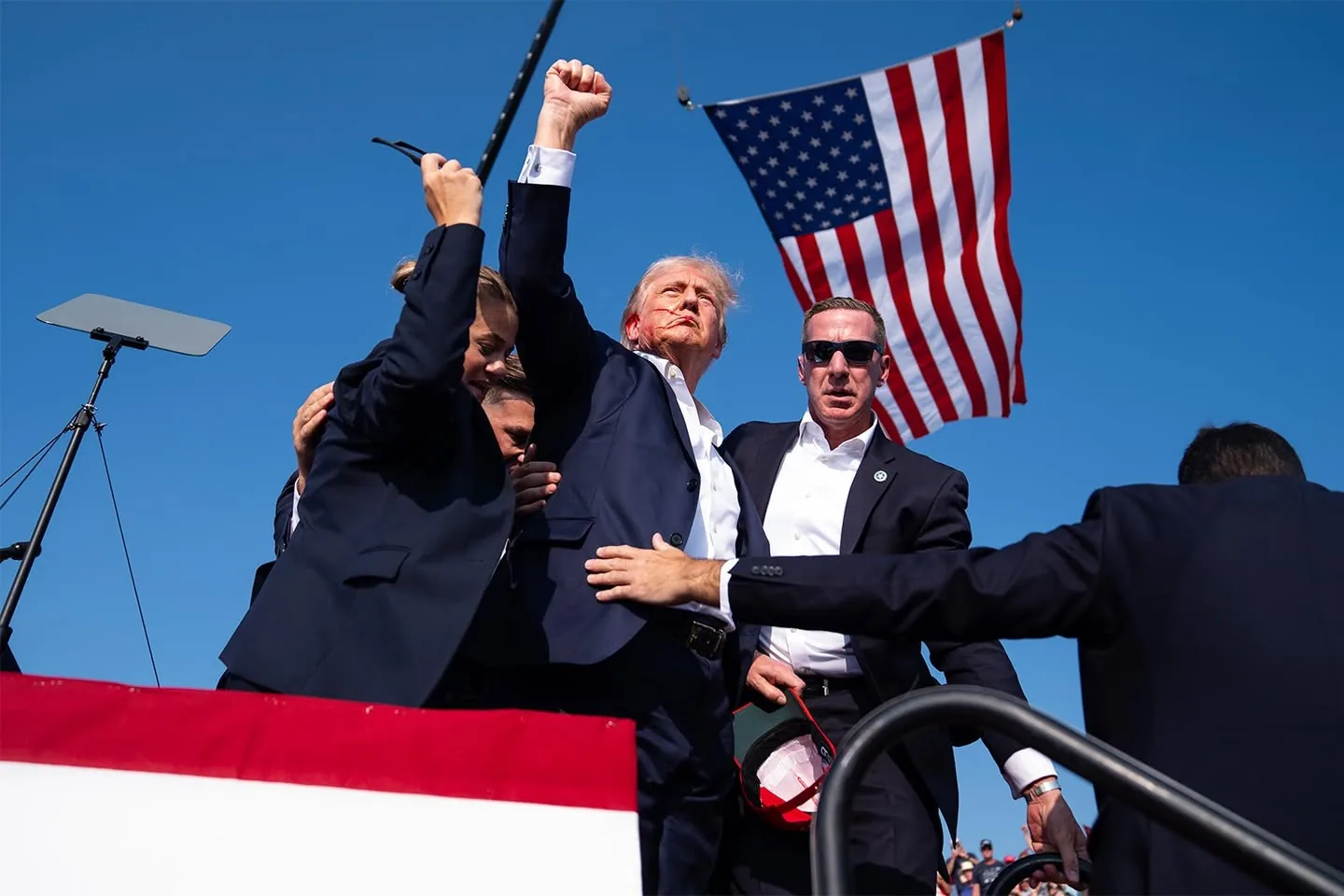 Donald Trump is at an outdoor event, surrounded by people. He is raising his fist in the air with a determined expression. Trump is wearing a dark suit with a white shirt, and is positioned prominently in the center of the group. To his side is Sean Curran, wearing sunglasses and a dark suit, who appears to be part of his security detail. Curran is holding a hat with red accents. The scene is set against a clear blue sky, with an American flag prominently displayed in the background, adding a patriotic element to the scene. There is a podium with a teleprompter to the left, suggesting a speaking engagement or rally atmosphere. The mood is energetic and assertive, with the crowd's presence implied by the composition. The image captures a dynamic moment of interaction and engagement, emphasized by the vivid colors and strong gestures.