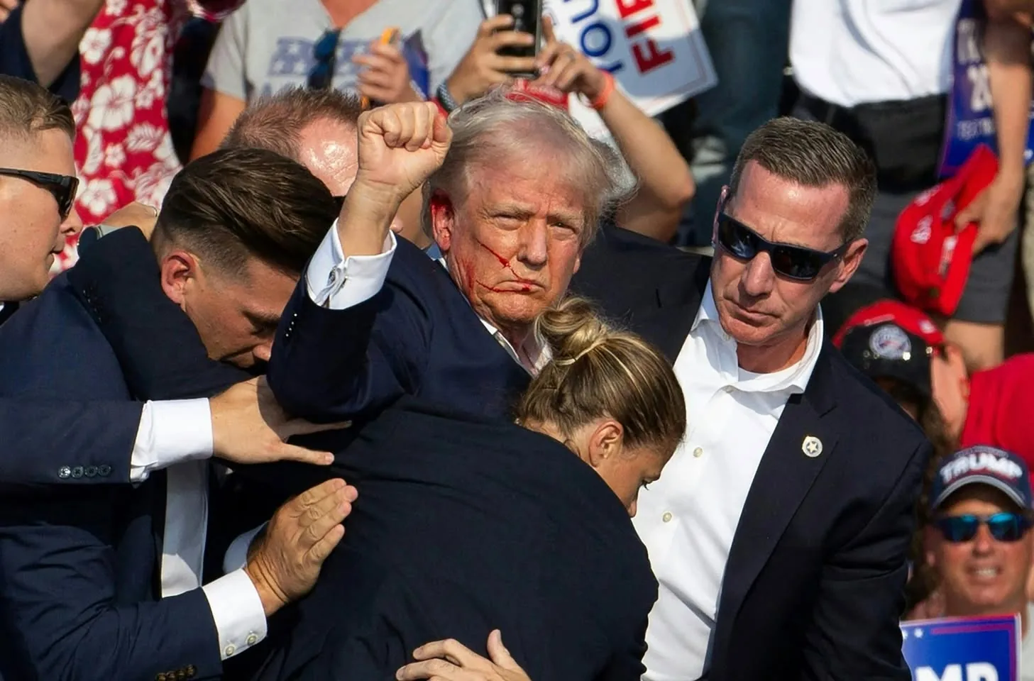 Donald Trump is at the center of the image, surrounded by security personnel, including Sean Curran. Donald Trump has a stern expression, with visible red marks on his face, suggesting an injury or possibly a theatrical effect. His right arm is raised in a fist, a gesture often associated with defiance or solidarity. He is wearing a dark suit and white shirt, customary for formal events. Sean Curran and other individuals are wearing suits and sunglasses, indicating they are likely security personnel.
The background is filled with a dense crowd, some holding signs, including one that reads "You're Fired," reminiscent of Trump's catchphrase from his television career. The crowd is lively, with people capturing the moment on their phones. The lighting is bright and suggests it is daytime, likely at an outdoor political rally or event. The atmosphere appears charged with energy, typical of public appearances involving high-profile figures like Donald Trump.
