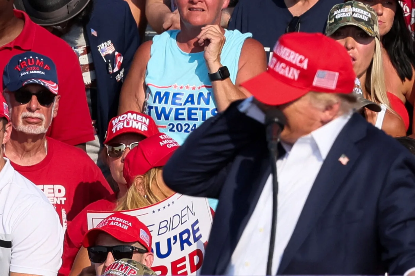 Donald Trump is featured prominently in this image, standing at a podium during what appears to be a rally. He is wearing a red "Make America Great Again" hat and a dark suit with a white shirt, partially obscured by the microphone. The crowd behind him is filled with supporters, many of whom are wearing Trump-themed hats and shirts, such as "TRUMP 2024 SAVE AMERICA" and "WOMEN FOR TRUMP". A notable sign in the background reads "BIDEN YOU'RE FIRED," indicating the political nature of the event. Another person is wearing a shirt with "MEAN TWEETS 2024" printed on it, referencing Trump's previous activity on social media. The mood of the crowd seems enthusiastic and supportive, with vibrant colors like red, white, and blue dominating the scene. The lighting is bright and suggests a daytime outdoor setting, contributing to the energetic atmosphere typical of political rallies.