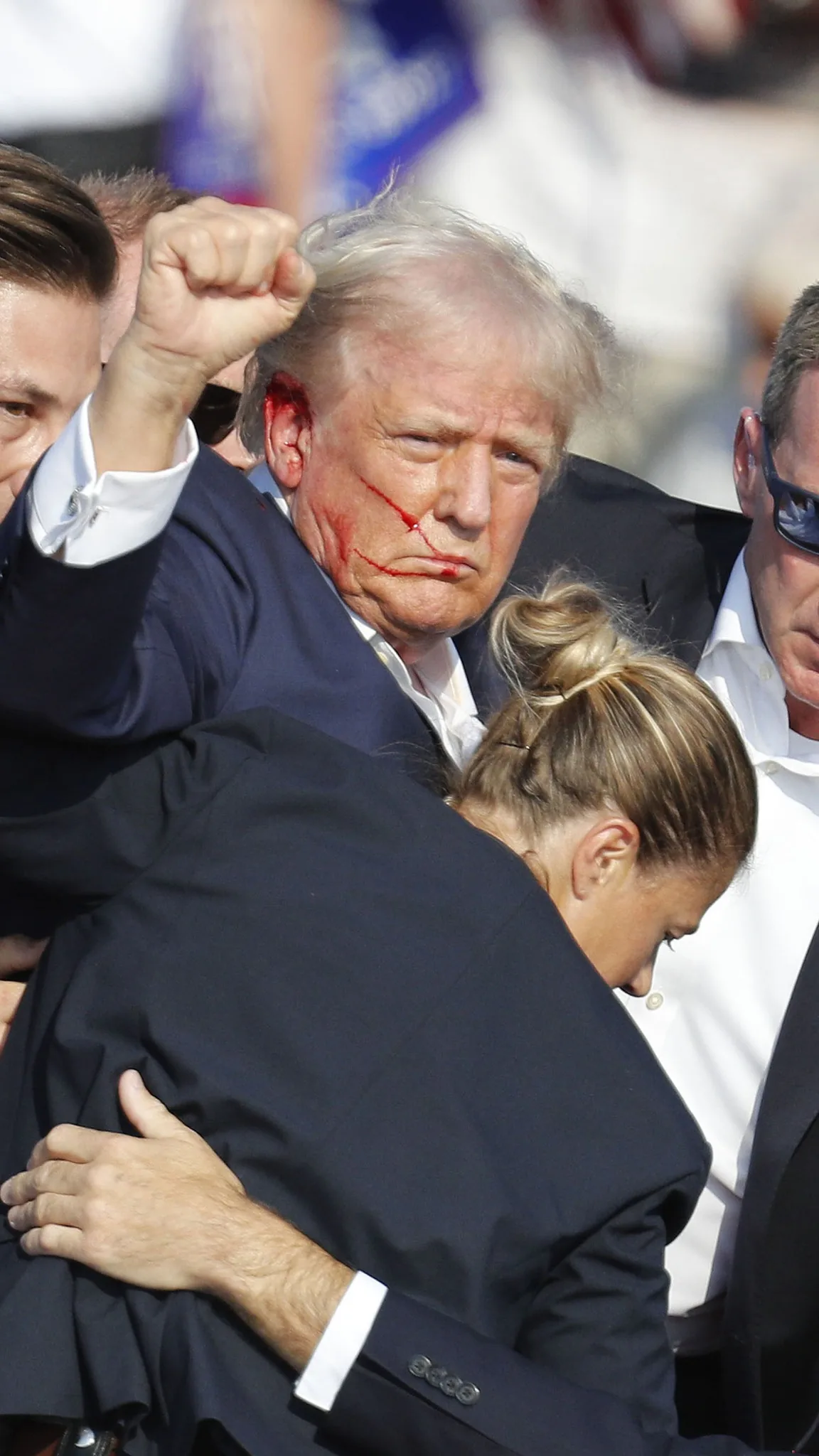 The image shows Donald Trump in an outdoor setting surrounded by several people, including what appears to be security personnel. Trump is in the center of the image, wearing a dark suit and white shirt. He has a serious expression, and a noticeable red mark is visible on his right cheek, resembling a fresh wound or fake blood. His right hand is raised in a fist, a gesture that can imply defiance or solidarity.
The background is blurred, indicating a crowd or public event, with various individuals partially visible. The lighting is bright, typical of a daytime event, suggesting the scene takes place outdoors during the day. The attire of those surrounding Trump is formal, with suits and ties, fitting for an official or public occasion. The focus on Trump, combined with the gestures and the presence of security, conveys a sense of urgency or a significant moment.