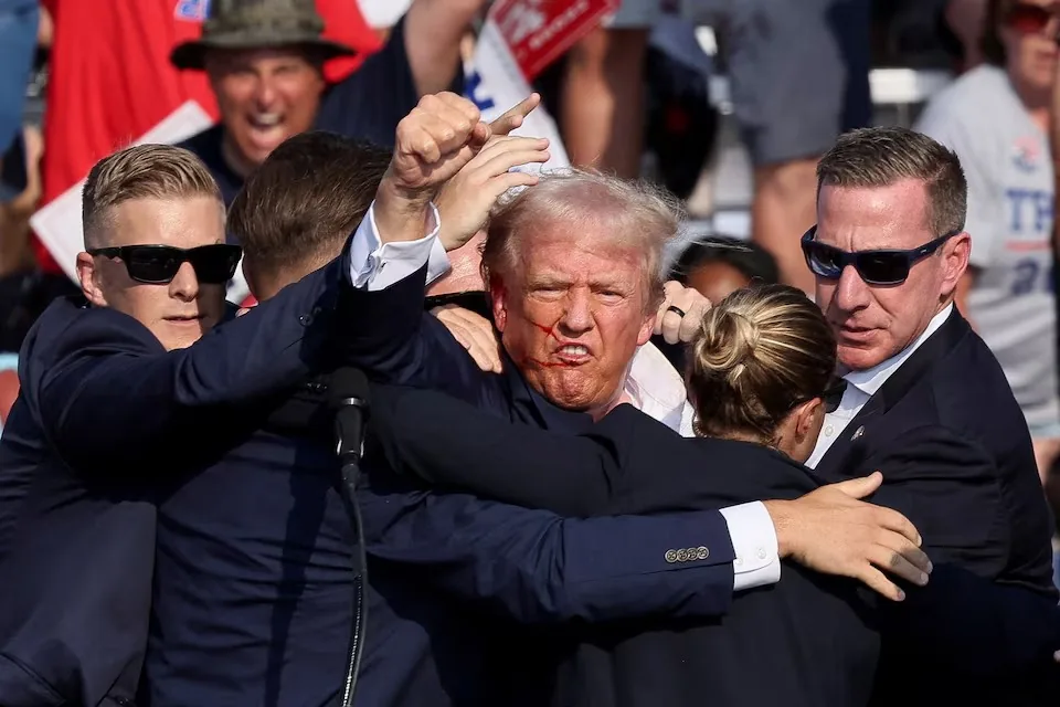 Donald Trump is at the center of this dynamic scene, surrounded by several individuals, likely security personnel, indicating a heightened sense of urgency and protection. Sean Curran is also named, though it's unclear from the image who he is specifically, as multiple people are present. Trump's expression appears tense or strained, with a slight frown and puckered lips, possibly indicating stress or a reaction to an external stimulus. The security personnel are dressed in formal suits, wearing sunglasses, and are closely huddled around him, some with their arms extended, possibly to shield or guide him.
The setting seems to be outdoors, possibly at a public event or rally, as evidenced by the presence of a cheering crowd in the background, some holding signs or banners. The lighting is bright and suggests a sunny day, casting clear shadows and highlighting the focused determination on the faces of the security team. The composition centers around the protective huddle, emphasizing the seriousness of the situation. The crowd in the background contributes to the charged atmosphere, with their expressions and gestures varied but animated.