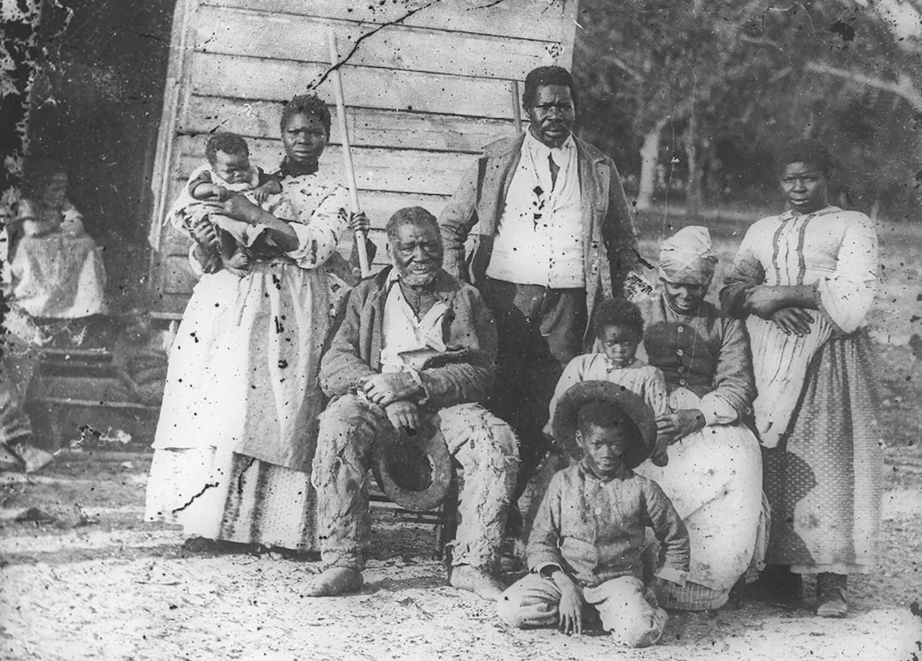The image is a historical black-and-white photograph depicting a group of individuals outdoors, likely from the 19th century. The group consists of several adults and children, positioned in front of a wooden structure. An older man is seated in the center, holding a hat, and he appears to be the focal point of the photograph. His expression is relaxed as he gazes at the camera.
Surrounding him are women and men, some standing and some seated, with one woman holding a baby in her arms. The individuals are dressed in clothing typical of the era, with simple, utilitarian garments. The women wear long dresses, and one has a headscarf tied around her head. The children, one of whom is seated on the ground wearing a hat, appear to be looking curiously at the camera.
The setting is outdoors, with the wooden structure providing a rustic backdrop, and trees can be seen in the background, suggesting a rural environment. The photograph is marked with age, featuring scratches and imperfections that add to its historical authenticity. The lighting appears natural, highlighting the details in the subjects' clothing and expressions.