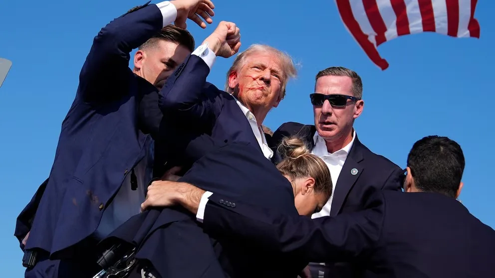 Donald Trump is at the center of the image, surrounded by several security personnel. He is looking upwards, with a serious expression, while being closely guarded by individuals in dark suits. Sean Curran is among the security team, recognizable by his sunglasses and attentive posture. The group appears to be moving or responding to a situation, with one guard reaching forward protectively. An American flag is visible in the background, fluttering against a clear blue sky, adding a patriotic element to the scene. The lighting is bright, suggesting it is daytime, and the overall mood is tense and vigilant, indicative of a public event or rally where security is paramount. The composition captures a moment of action and urgency, with all eyes on Donald Trump.