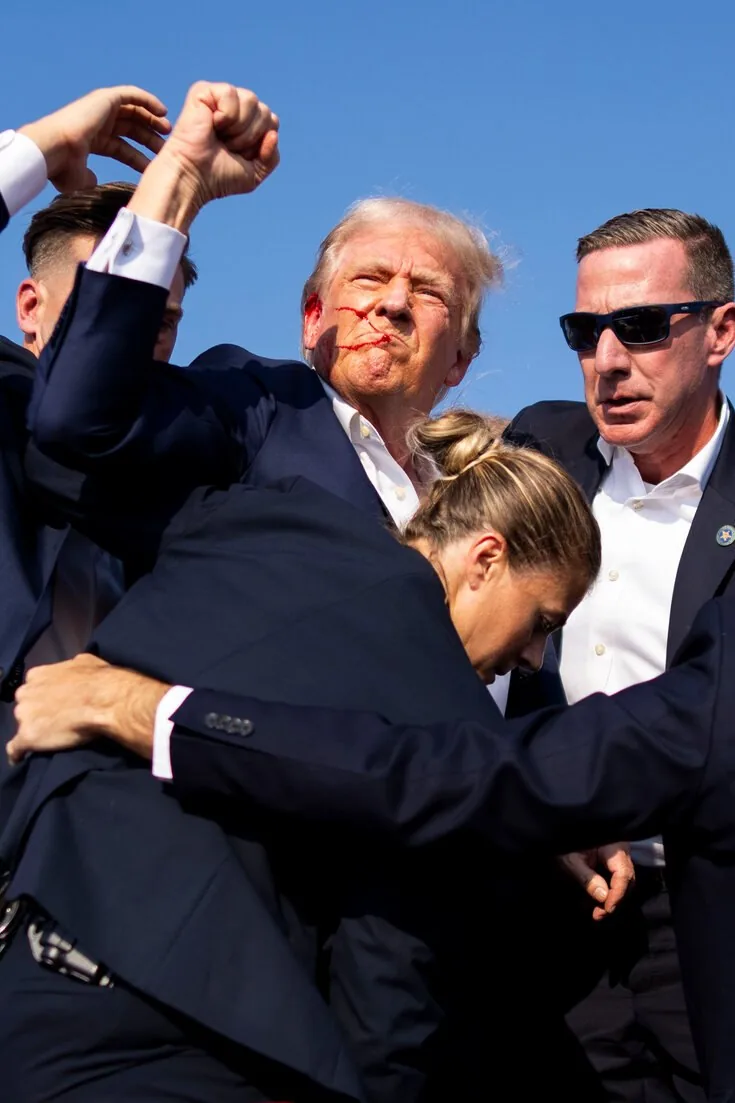 Donald Trump is prominently featured in this outdoor scene under a clear blue sky. He is surrounded by a group of people, one of whom is identified as Sean Curran. Trump is being supported by several security personnel, indicating a protective or urgent situation. He appears to have some red marks on his face, suggesting an incident or event that might have caused them. Dressed in a dark suit with a white shirt, Trump is captured with a determined expression, as if trying to maintain composure or address the crowd. Sean Curran, among the others, is also wearing a dark suit and sunglasses, projecting a professional security presence. The mood of the image seems tense, with the focus on ensuring Trump's safety. The bright lighting highlights the scene, emphasizing the seriousness of the moment, with no visible text or branding in the image.