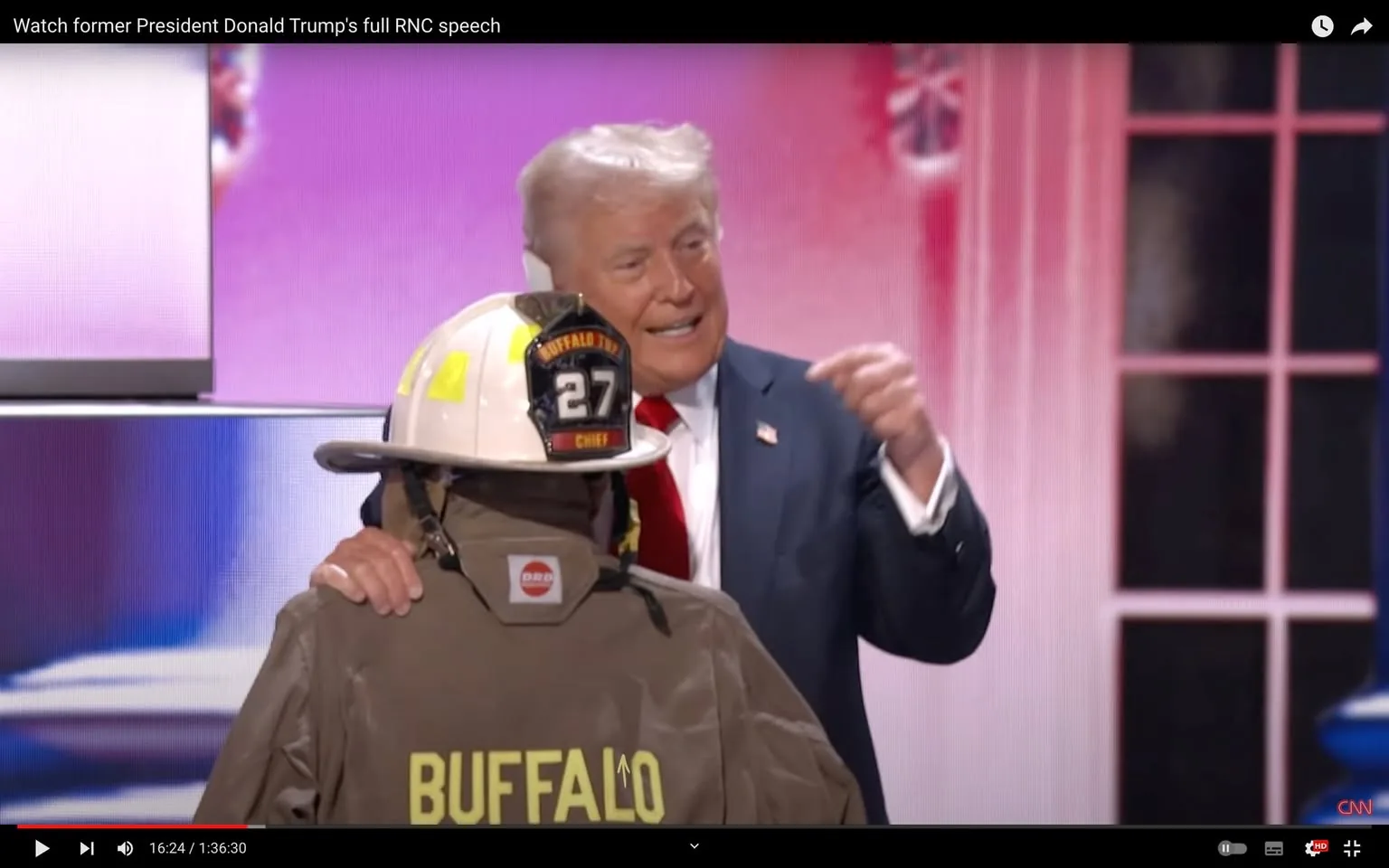 In this image, Donald Trump is on stage, likely during a public event or speech, as indicated by the surrounding setting. He is positioned to the right, wearing a dark suit with a white shirt and a bright red tie, characteristic of his style during official appearances. His expression appears animated, and he seems to be speaking or gesturing energetically towards the audience or someone on stage with him.
In the foreground, a firefighter in full gear stands with their back to the camera, prominently displaying the word "BUFFALO" on their jacket. The firefighter's helmet is marked with "27" and "CHIEF," suggesting a leadership role within their unit. The juxtaposition of Donald Trump and the firefighter creates a striking scene, likely symbolic or ceremonial.
The backdrop is a vibrant mix of reds and pinks, possibly reflecting a patriotic or thematic display. The image is a snapshot from a video titled "Watch former President Donald Trump's full RNC speech," as seen on YouTube, and it is published by CNN. The timestamp at the bottom indicates the video is 16 minutes and 24 seconds into its total duration of 1 hour and 36 minutes.