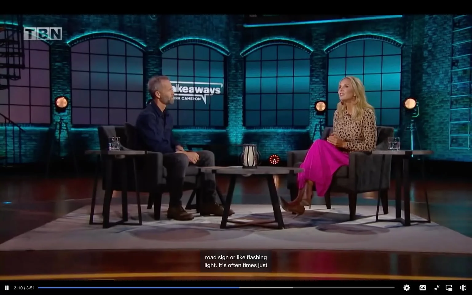 In this scene from the talk show "Takeaways with Kirk Cameron," Elisabeth Hasselbeck is engaged in an interview setting. The studio features a modern and stylish design, with large, arched windows in the background and a cool blue lighting scheme that creates a relaxed and inviting atmosphere. Elisabeth Hasselbeck is seated on the right, wearing a leopard-print top and a vibrant pink skirt, paired with brown boots, giving her a fashionable appearance. The host, Kirk Cameron, sits to the left, dressed in a dark shirt and gray pants. Both are seated in dark upholstered chairs with a small wooden table between them, holding a decorative vase and glasses of water. The TBN logo is visible in the upper corner, denoting the network, and the show's title is displayed prominently behind them. The conversation is captured with subtitles, reflecting a thoughtful discussion. The overall mood is professional yet casual, typical of talk show formats.
- Title: "Takeaways with Kirk Cameron"
- Site / app: "TBN"
- Captions / subtitles: "road sign or like flashing light. It's often times just"