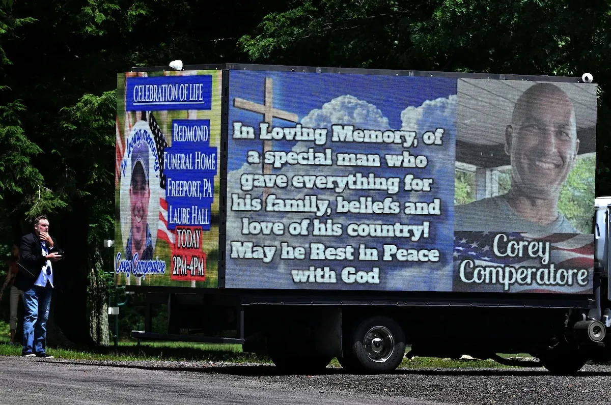 ```
**SECTION 2: Rich Prose Description**
The image shows a large digital sign vehicle set up in a lush, green outdoor setting. The sign is part of a "Celebration of Life" event for Corey Comperatore, prominently displayed on the side of the vehicle. The left panel of the sign includes a photograph of Corey wearing a cap, with text providing details of the memorial service: "REDMOND FUNERAL HOME, FREEPORT, PA, LAUBE HALL, TODAY 2PM-4PM." The background features an American flag motif, reflecting patriotic themes.
The right panel features a larger, smiling photo of Corey Comperatore against a backdrop of clouds and a wooden cross, symbolizing peace and remembrance. The text honors him as a "special man who gave everything for his family, beliefs, and love of his country," and includes a message of rest with God. The setting suggests a somber yet celebratory mood, with the digital display providing a modern touch to traditional memorial practices.
In the foreground, a man stands with a contemplative expression, possibly a guest or organizer, holding a phone. He is dressed in casual attire, indicating a respectful yet relaxed event atmosphere. The overall scene conveys a heartfelt tribute, blending personal remembrance with public acknowledgment.