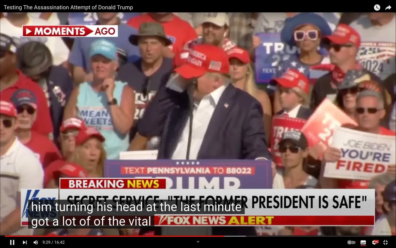 The image is a video frame from a YouTube video titled "Testing The Assassination Attempt of Donald Trump." It appears to capture a moment during a political rally involving Donald Trump. In the foreground, Donald Trump is wearing a red "Make America Great Again" cap and a dark suit, standing at a podium with a sign that reads "TEXT PENNSYLVANIA TO 88022." He seems to be adjusting something on his head or wiping his brow, partially obscured by his hand. Behind him, a crowd of supporters is visible, many wearing red caps, sunglasses, and patriotic attire. Some hold signs supporting Trump and expressing disdain for his political opponents, visible in slogans such as "Joe Biden You're Fired."
Visible on-screen text includes a "BREAKING NEWS" banner from Fox News, indicating that the Secret Service has stated, "THE FORMER PRESIDENT IS SAFE." The scene is set outdoors during the day, with bright lighting suggesting a sunny day. The mood appears tense, possibly due to the context of the news banner. The frame includes subtitles discussing a critical moment involving Trump turning his head.