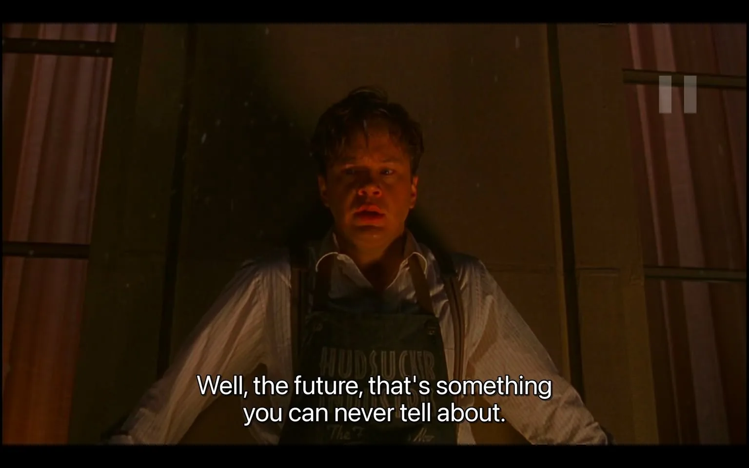 The image is a screenshot from the film "The Hudsucker Proxy." It features a man leaning back against a wall, his expression appearing contemplative or resigned. The lighting is dramatic, casting shadows on his face and highlighting the intensity of the moment. He is dressed in a white shirt with suspenders and a dark apron bearing the word "HUDSUCKER" partially visible. This suggests his role or affiliation with a business or company, which ties into the film's setting and plot. The background is dimly lit, possibly indicating an indoor setting with a somber or tense atmosphere. The overall composition draws attention to the character's emotional state, enhancing the scene's mood. Subtitles at the bottom read, "Well, the future, that's something you can never tell about," adding depth to the character's introspection.