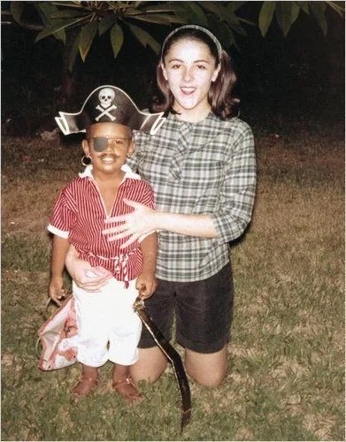 The image captures a moment from the past featuring "Infant Barack Obama" and a woman outdoors at night. The young child is dressed in an adorable pirate costume, complete with a hat adorned with a skull and crossbones, an eye patch, a red and white striped shirt, white pants, and a toy sword. His expression is joyful, with a broad smile, radiating a sense of playfulness and excitement typical of a child's imagination during dress-up. Standing beside him is a woman wearing a checked shirt and dark shorts, embracing the child affectionately to ensure he is steady and comfortable. The background is a grassy area with some tree foliage visible overhead, suggesting they are in a garden or park setting. The lighting appears to be artificial, possibly from a camera flash, highlighting the subjects against the dim surroundings. The photograph has a nostalgic feel, capturing a playful and tender family moment.