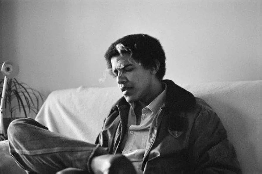 Barack Obama is captured in a candid black-and-white photograph, seated casually on a couch. The setting suggests a relaxed, informal environment, possibly a living room. Obama is dressed in a collared shirt and a jacket, exuding a laid-back demeanor. His posture is informal, with one leg crossed over the other. His expression appears contemplative or reflective, as he seems to be deep in thought or engaged in conversation. The background includes a lamp and some plants, adding to the cozy, home-like atmosphere. The lighting is soft, typical of natural indoor light, creating gentle shadows and highlighting his features subtly. The image has a nostalgic feel, likely taken during his younger years, offering a glimpse into a quieter, personal moment.