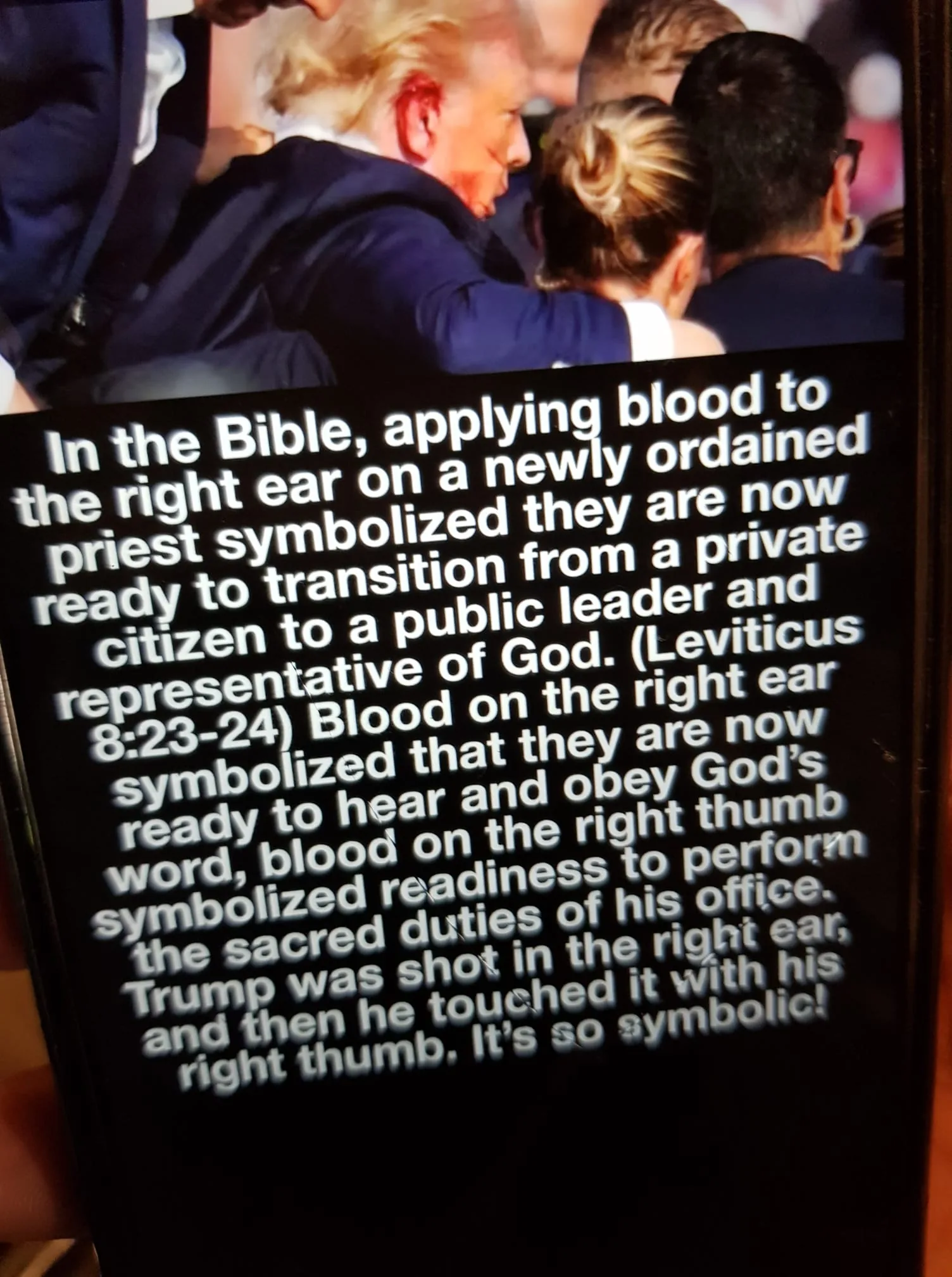 The image shows a man, identified in the hint text as Trump, surrounded by several people, suggesting he is at a public event or gathering. The focus is on his right ear, which appears to have a red mark, resembling blood. The crowd around him includes people with different hair colors and styles, hinting at a diverse group. The setting seems to be indoors, possibly at a rally or ceremony, indicated by the close proximity and the formal attire of the individuals.
The lighting is bright, highlighting the central figure, while the background remains slightly out of focus. The mood appears serious or intense, possibly due to the context provided in the text. The text is prominently displayed in white against a dark background, explaining a symbolic interpretation of blood application in the Bible and relating it to the scene involving Trump, suggesting a transition to a public leadership role. The text references Leviticus 8:23-24, symbolizing readiness to hear and obey God's word and perform sacred duties, drawing a parallel to the depicted scene.