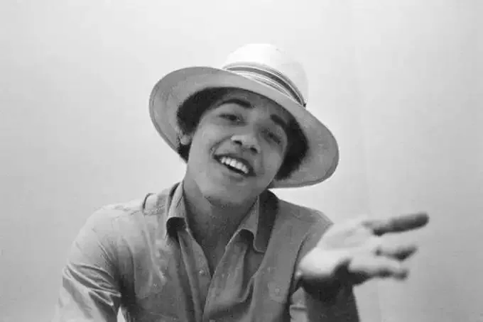 This black and white photograph captures a young Barack Obama in a relaxed and informal pose. He is seated indoors against a plain background, which emphasizes his presence in the frame. Barack Obama is wearing a light-colored button-up shirt, and he has a straw hat with a dark band adorning his head, adding a touch of casual style to the portrait. His expression is engaging and friendly, with a wide, open smile that exudes a sense of youthful exuberance and approachability. His left hand is slightly outstretched towards the camera, enhancing the sense of spontaneity and interaction with the viewer. The lighting is soft and even, typical of indoor settings, which helps in highlighting the subject's features without casting harsh shadows. The photograph, likely taken during his early years, offers a glimpse into the personal style and charisma that would later become synonymous with his public persona.