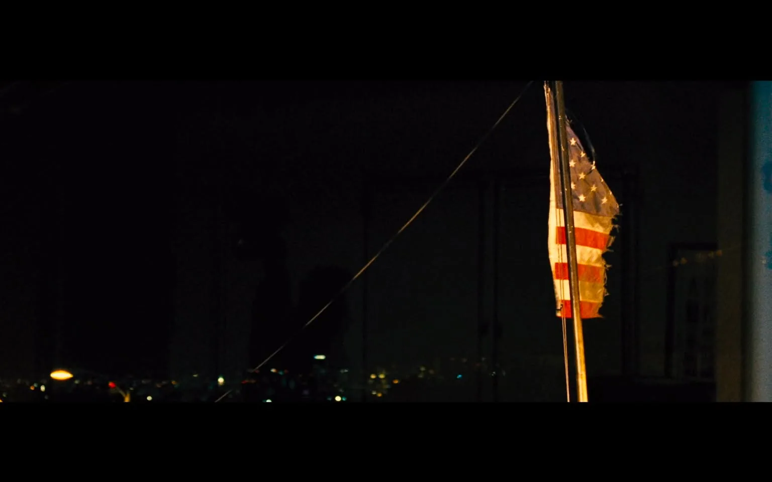 The image depicts an indoor nighttime scene where a U.S. flag is prominently featured. The flag is positioned on the right side, slightly tattered and illuminated by a warm light source. Its colors are slightly muted, but the stars and stripes are clearly visible. The background is dark, suggesting nighttime, with faint city lights visible outside a window, providing a contrast to the flag's brightness. The setting feels quiet and somewhat somber, with the flag standing as the main focal point. There are no people or noticeable activities in the frame, contributing to a still, contemplative atmosphere. This composition evokes a sense of patriotism mixed with nostalgia or reflection. The overall color palette is dark with occasional bright spots from the city lights and the flag's illumination.