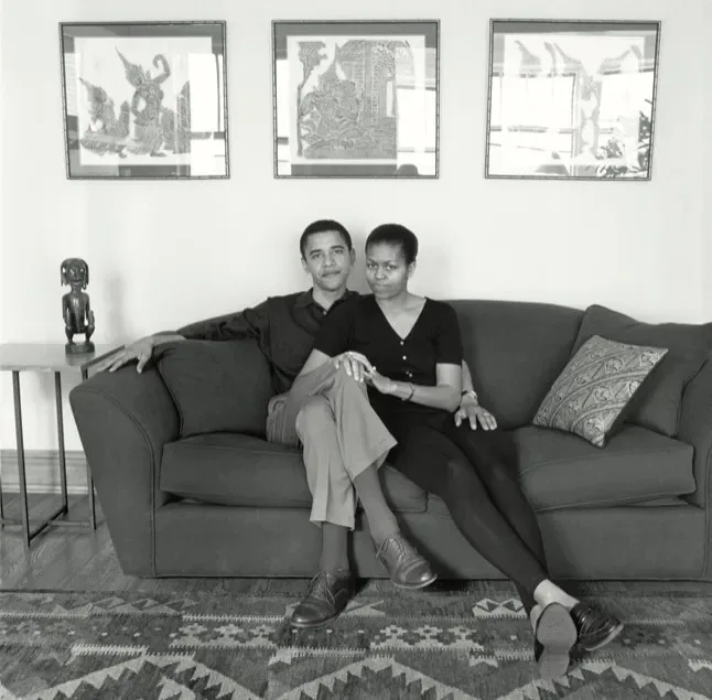 Barack Obama and Michelle Obama are seated together on a couch in a cozy living room setting. The image is in black and white, giving it a classic and timeless feel. Barack Obama, dressed in a dark shirt and light-colored pants, has a relaxed demeanor, one arm draped over the back of the couch. Michelle Obama is leaning slightly towards him, wearing a dark top and pants, her expression calm and composed. They are positioned closely, suggesting intimacy and comfort. Behind them, three framed pieces of artwork add an aesthetic touch to the room, and a small table beside the couch holds a decorative statue. A patterned rug covers the wooden floor, contributing to the warm atmosphere of the room. The overall mood is one of calmness and togetherness, capturing a personal moment between the couple.