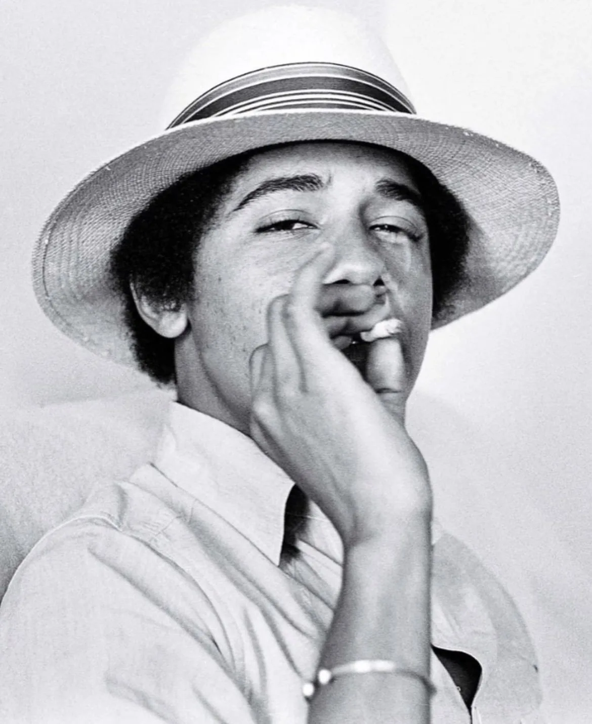 Barack Obama is captured in a candid, black-and-white photograph. He is wearing a light-colored shirt and a stylish straw hat with a band. The image exudes a relaxed and laid-back vibe, with Barack Obama looking directly at the camera with a casual expression. He is holding a cigarette to his mouth, which adds to the informal and youthful atmosphere of the picture. The lighting is soft, enhancing the nostalgic and intimate feel of the scene. The close-up composition focuses on his face, making his expression the central element of the photograph. The choice of monochrome adds a timeless quality to the image, highlighting texture and contrast. Overall, the photograph is a snapshot of a youthful moment, capturing a sense of ease and nonchalance.