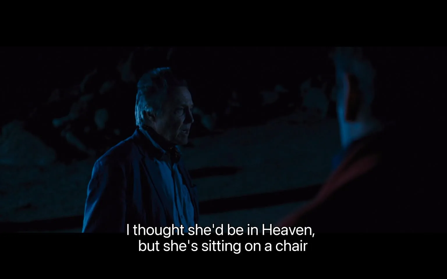 1) Rich description: The scene is set at night, outdoors, with dim lighting that casts a bluish hue over the surroundings. One man is visible in the frame, standing to the left, partially illuminated by the faint light. His expression appears thoughtful or introspective. He is wearing a dark jacket and shirt, blending subtly with the night. The background is shrouded in darkness, with indistinct shapes suggesting rocks or uneven terrain, adding to the mysterious and somber ambiance. The conversation seems to carry an emotional weight, highlighted by the caption, "I thought she'd be in Heaven, but she's sitting on a chair," suggesting a poignant or ironic reflection. The framing focuses on the older man, with another figure visible in silhouette on the right, perhaps listening or responding silently.
2) On-screen text block:
- Captions / subtitles: "I thought she'd be in Heaven, but she's sitting on a chair"