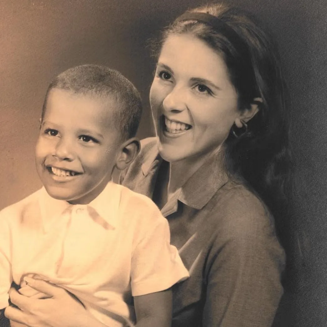 In this warm and intimate portrait, Baby Barack Obama is pictured with his mother in a classic studio setting. The photograph is in sepia tones, giving it a timeless, nostalgic quality. Baby Barack Obama, a young boy in this image, is wearing a light-colored, button-up shirt and looks cheerful, smiling brightly at something beyond the frame. His mother, sitting close beside him, is wearing a collared shirt and a headband that keeps her hair elegantly in place. Her expression is joyful and affectionate, her eyes crinkling with a smile as she gazes alongside her son. The composition captures a tender moment of familial bond, with soft lighting enhancing the warmth and closeness shared between the two. The background is simple and non-distracting, focusing the viewer’s attention entirely on the subjects’ expressions and connection.