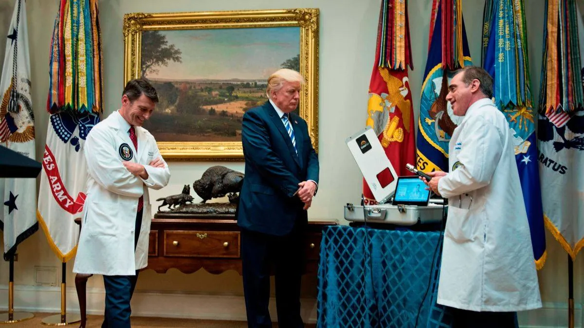 In this image, Donald Trump is standing in the center of a room, flanked by two individuals wearing white medical coats, suggesting a formal presentation or meeting. The setting appears to be an official room, as indicated by the presence of multiple military flags and a painting on the wall. Donald Trump is dressed in a dark suit with a striped tie, standing with his hands clasped in front of him, exuding a formal demeanor.
The two individuals in white coats are likely medical professionals, with one holding a device or remote, possibly for a demonstration. Both have attentive and engaged expressions. A table draped with a blue cloth holds a piece of equipment, which might be part of the demonstration. The room's decor includes a framed landscape painting and a buffalo sculpture on a wooden sideboard, adding a sense of elegance to the scene.
The lighting is bright and even, illuminating the details of the room and the participants. The overall mood of the image is serious yet cordial, reflecting a professional engagement.