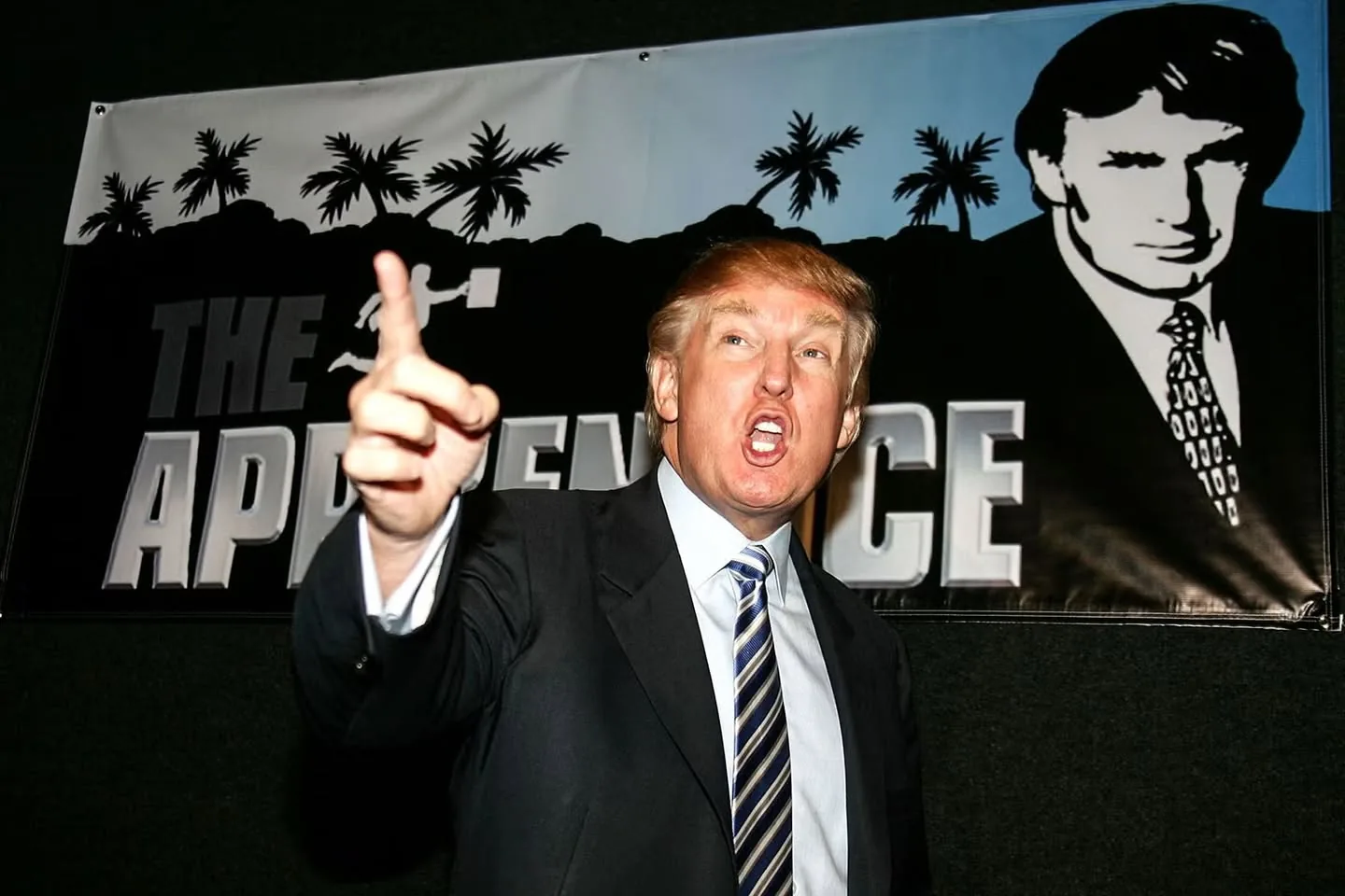 Donald Trump is captured in a dynamic moment, speaking energetically at an event. He is wearing a dark suit paired with a striped tie, and his expression is animated, with his mouth open as if he is emphasizing a point. His right hand is raised, index finger pointing upward, adding to the intensity of his speech. Behind him is a banner featuring the text "THE APPRENTICE," along with a stylized black-and-white image of Donald Trump. The banner also depicts palm trees silhouetted against a blue background, adding a tropical flair to the scene. The lighting is bright and focused on Donald Trump, accentuating his presence against the dark background. This setting suggests a promotional appearance or a press event for the television show "The Apprentice." The mood is assertive and commanding, reflecting Donald Trump's public persona during his time on the show.