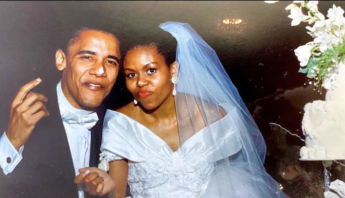 In this image, we see Barack Obama and Michelle Obama on their wedding day. The scene captures a joyful and intimate moment between the couple. Barack Obama is dressed in a classic black tuxedo with a white bow tie and shirt, exuding elegance and charm. Beside him, Michelle Obama looks radiant in a beautiful white wedding gown adorned with intricate lace patterns. Her dress has an off-the-shoulder design, complemented by a long, flowing veil. Her earrings are large and elegant, adding to her bridal look. The couple is posing closely together, with Barack Obama making a playful gesture with his hand. The background is softly lit, and there is a slight blur, focusing the attention on the couple. To the side, part of a wedding cake adorned with flowers is visible, emphasizing the celebratory atmosphere. The overall mood is one of happiness and celebration, capturing a cherished moment in their lives.