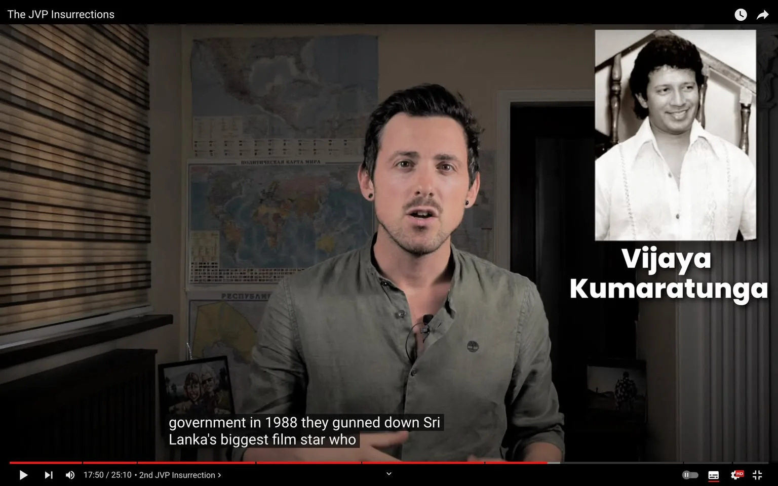 1) Rich description:
The video frame is from a segment titled "The JVP Insurrections." It features a presenter in the foreground, positioned to the right, wearing a muted green shirt with a small logo on the chest. He appears to be speaking, with a focused expression, possibly explaining or narrating the content. The setting is indoors with a warm color palette, and behind him are maps of the world, indicating a historical or educational theme.
In the upper right corner, there is a black-and-white photograph of Vijaya Kumartunga, accompanied by his name in bold white text. This suggests that the segment is discussing events related to him, likely his impact or involvement in the historical context being addressed.
The room is softly lit, and the background includes window blinds, giving a sense of a study or office environment. The subtitles at the bottom mention a significant event from 1988, adding historical depth to the discussion.
2) On-screen text block:
- Title: "The JVP Insurrections"
- Captions / subtitles: "government in 1988 they gunned down Sri Lanka's biggest film star who"
