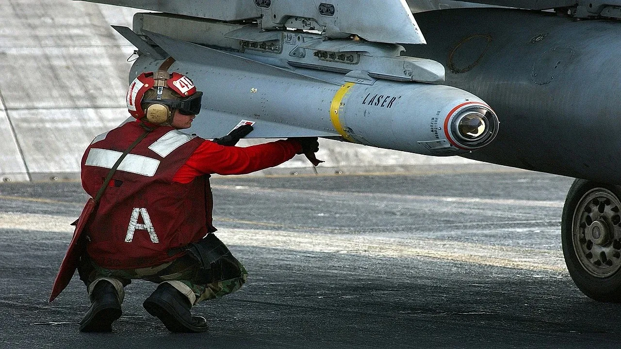 A military personnel is seen kneeling on an aircraft carrier deck, working on an aircraft. The person is wearing a red vest with reflective white stripes and the letter "A" printed on the back, along with a helmet and protective goggles. The individual appears to be involved in maintenance or inspection tasks, holding onto a missile attached to the aircraft's wing. The missile is marked with the word "LASER", indicating it is likely a laser-guided munition.
The aircraft's landing gear and part of the wing are visible, with the scene taking place during the day under bright lighting conditions. The surface of the deck appears to be metallic and slightly worn, typical of an operational aircraft carrier. The colors are primarily muted greys and reds, reflecting the utilitarian nature of the setting. The overall mood is one of focus and precision, highlighting the disciplined environment of military operations.