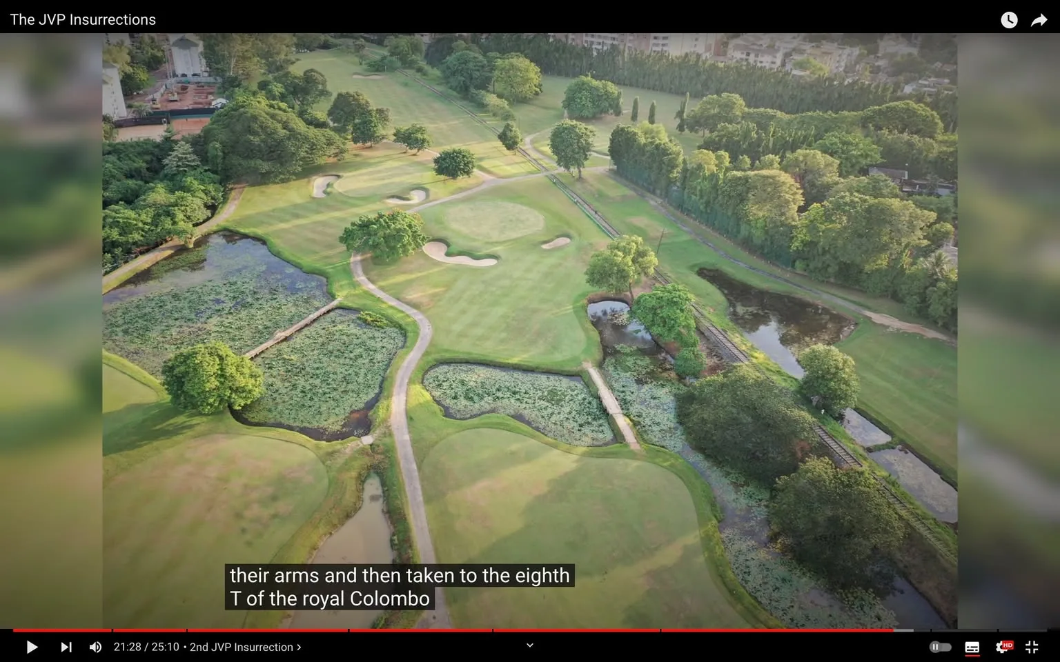 ```
The image is an aerial view of a golf course, possibly part of the Royal Colombo Golf Club, as mentioned in the subtitles. The landscape stretches across the frame, showcasing well-maintained green fairways, sand bunkers, and surrounding patches of trees and water bodies. The paths meander through the course, providing a neat and organized look. The lighting suggests daytime, with sunlight casting soft shadows from the trees. The terrain appears lush and vibrant, indicating a location that is well-cared for. In the background, there are hints of buildings, bordered by a dense line of trees that add to the sense of seclusion and tranquility typical of a golf course setting. The video seems to be discussing the JVP Insurrections, a significant historical event in Sri Lanka, with the caption related to activities at the eighth hole of the golf course. The video is titled "The JVP Insurrections," which indicates its focus on historical or political content. The medium is a video frame, and the language of the subtitles is English.