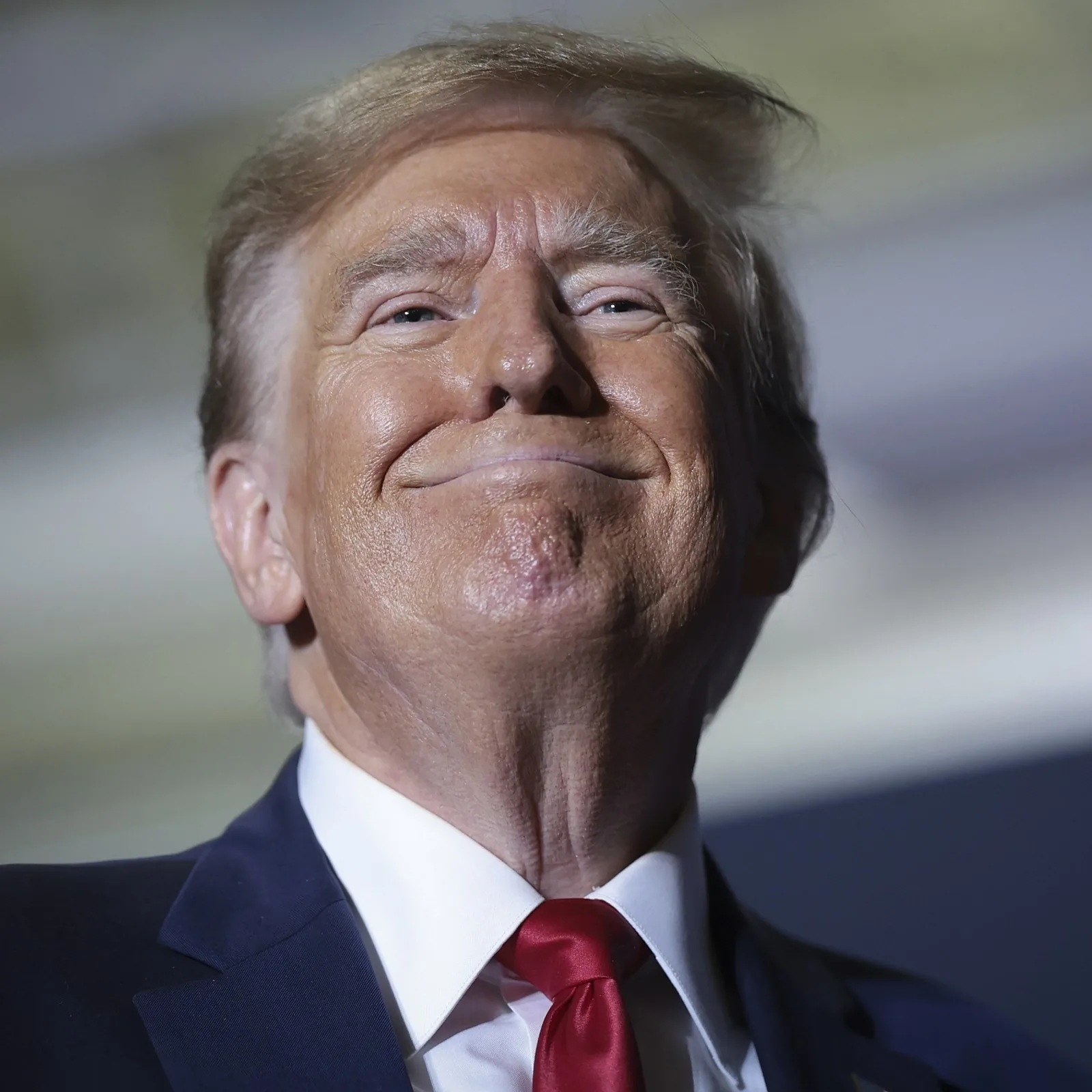 Donald Trump is featured in this close-up image, where he appears to be indoors, possibly at a public event or speech. His expression is slightly smiling, with a hint of a smirk. He is wearing a dark blue suit, a crisp white shirt, and a bright red tie, which are characteristic elements of his typical public attire. The lighting is soft, emphasizing his facial features and casting gentle shadows that add depth to the image. The background is blurred, focusing attention entirely on Donald Trump, making him the central subject. The image captures a moment that suggests confidence or contemplation. The angle is slightly upward, giving a sense of prominence or authority to the figure. The image quality is high, with clear details and vibrant colors, typical of professional photography at significant events.