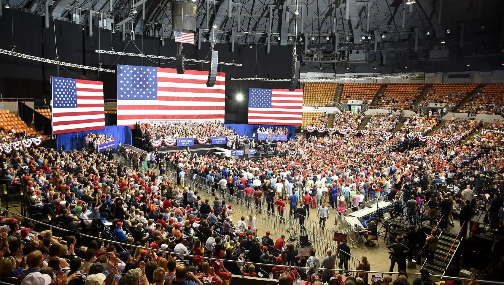 The image captures a large indoor political rally with a massive audience seated and standing in an arena. The setting is vibrant with patriotic decorations, prominently featuring two large American flags displayed on the stage backdrop. The audience, dressed casually, includes many wearing red hats, suggesting a cohesive group supporting a specific cause or candidate. A prominent stage is visible where speakers are likely addressing the crowd, marked by the phrases "PROMISES MADE" and "PROMISES KEPT," indicating the event's agenda or theme.
The lighting is bright, illuminating the entire venue, which is filled with rows of seats and standing areas. People appear engaged and attentive, with some taking photos or recording the event. The arena's structure is modern, with a high ceiling and visible lighting rigs. Attendees are seated in sections, with some areas cordoned off, possibly for media or security personnel. The atmosphere is lively, suggesting a significant and organized gathering, likely for a political campaign or support rally.