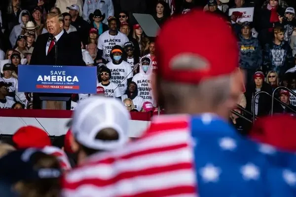 Donald Trump is delivering a speech at a political rally. He stands at a podium with a sign that reads "SAVE AMERICA," addressing a large crowd. Trump is dressed in a dark suit and red tie, and he appears focused and engaged. In the audience, people are wearing a variety of hats, including some with patterns resembling the American flag, indicating a strong patriotic theme.
Several attendees wear T-shirts with the phrase "BLACKS FOR TRUMP 2020," showing their support. The crowd is densely packed, with some individuals wearing cowboy hats, suggesting a diverse gathering. The overall atmosphere is lively and enthusiastic, with a sense of unity among the attendees. The lighting is bright, typical of an outdoor event, emphasizing the vibrant colors of the attire and signs. The image captures the dynamic interaction between Donald Trump and his supporters.