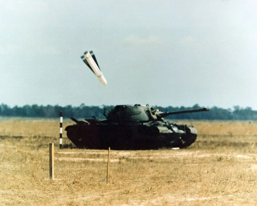The image captures a striking moment in a military setting, focusing on a tank situated in an open field. The tank appears stationary on the dry, grassy terrain, with a clear blue sky overhead. Above the tank, a munition is seen descending, suggesting an imminent impact. This dynamic scene conveys a sense of action and tension, typical of military exercises or demonstrations. The tank is painted in dark colors, blending with the muted tones of the landscape. In the foreground, small poles and markers are visible, likely used for navigational or boundary purposes during training. The lighting is natural, highlighting the details of both the tank and the dropping munition, while the background features a line of distant trees that frame the scene. The overall composition emphasizes the precision and intensity of modern military technology in action.