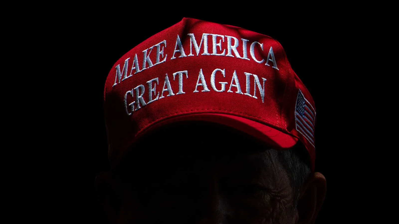 The image prominently features a red baseball cap with the slogan "MAKE AMERICA GREAT AGAIN" embroidered in white, capital letters. This phrase is widely associated with political campaigns in the United States. The cap is positioned against a dark, almost black background, which adds a dramatic contrast, making the bright red color of the cap stand out vividly. The embroidery is clear and centrally placed on the front of the cap, drawing immediate attention to the text. On the right side of the cap, a patch resembling the American flag is visible, adding a patriotic element to the visual. The lighting is focused on the cap, creating a sharp contrast with the surrounding darkness and emphasizing the text and colors. The image has a minimalistic and bold composition, with the hat serving as the sole focal point against the dark backdrop.