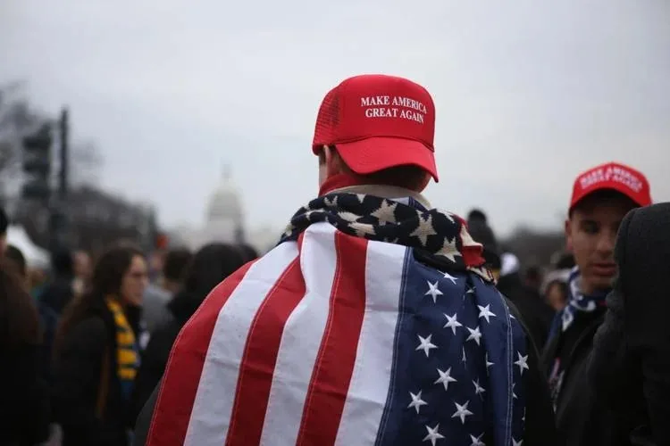 The image depicts a political rally, likely set in Washington D.C., as suggested by the visible dome of the Capitol building in the background. The central figure, seen from behind, is draped in an American flag and wearing a red "Make America Great Again" cap, a slogan associated with political campaigns in the United States. The crowd includes other individuals, some of whom are also wearing similar caps, indicating a shared political stance or support for the same cause or figure.
The mood of the scene appears serious and focused, with a cloudy sky that casts a muted light over the gathering. The individuals are dressed in winter attire, suggesting a cold day. The overall composition highlights the central figure's patriotic display, emphasizing the flag's colors and patterns, which stand out vividly against the more subdued tones of the crowd. This image captures the essence of a politically charged atmosphere, with symbols of national identity prominently displayed.