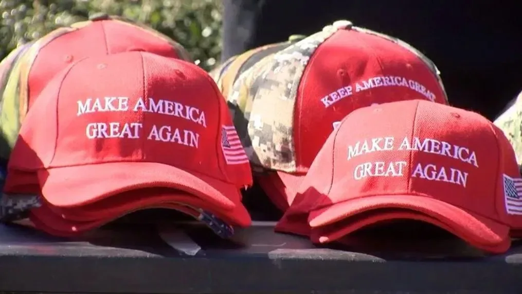 The image shows a collection of hats arranged on a surface. The dominant feature is the iconic red "Make America Great Again" caps, which have become synonymous with political support in the United States. The text is prominently embroidered in white across the front of the hats. Additionally, one of the hats displays the phrase "Keep America Great" in the background.
The hats are neatly stacked, with a camouflage-patterned cap visible behind the red ones, highlighting a variation in the collection. An American flag patch is sewn on the side of some caps, adding a patriotic element. The setting appears to be outdoors, indicated by the natural lighting and slight shadows. The image conveys a sense of political affiliation and merchandise display, typical in political rallies or events. The bright red color of the caps stands out distinctly against the neutral backdrop, drawing immediate attention to the text and theme.