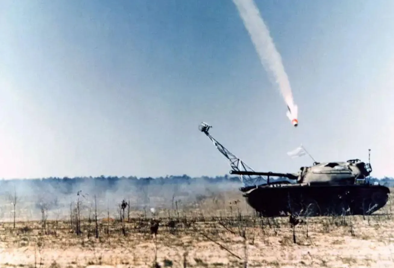 The image captures a dramatic outdoor scene featuring a tank positioned in a barren, desert-like landscape. The tank, with a camouflage pattern, is equipped with a distinctive arm extension mounted on its turret. From this arm, a missile appears to have been launched, with a visible trail of smoke against the clear blue sky. The missile is in mid-flight, heading upward at a steep angle, suggesting it has just been fired. The tank has a white flag attached to it, possibly indicating a training exercise or test rather than active combat. The foreground is sparse, with dry bushes scattered across the sandy ground. The scene is bathed in bright daylight, casting clear shadows and highlighting the movements and mechanisms involved in the missile launch. The setting suggests a military exercise area, emphasizing the technology and precision of modern warfare equipment. The overall mood is one of tension and anticipation, capturing a moment of action frozen in time.